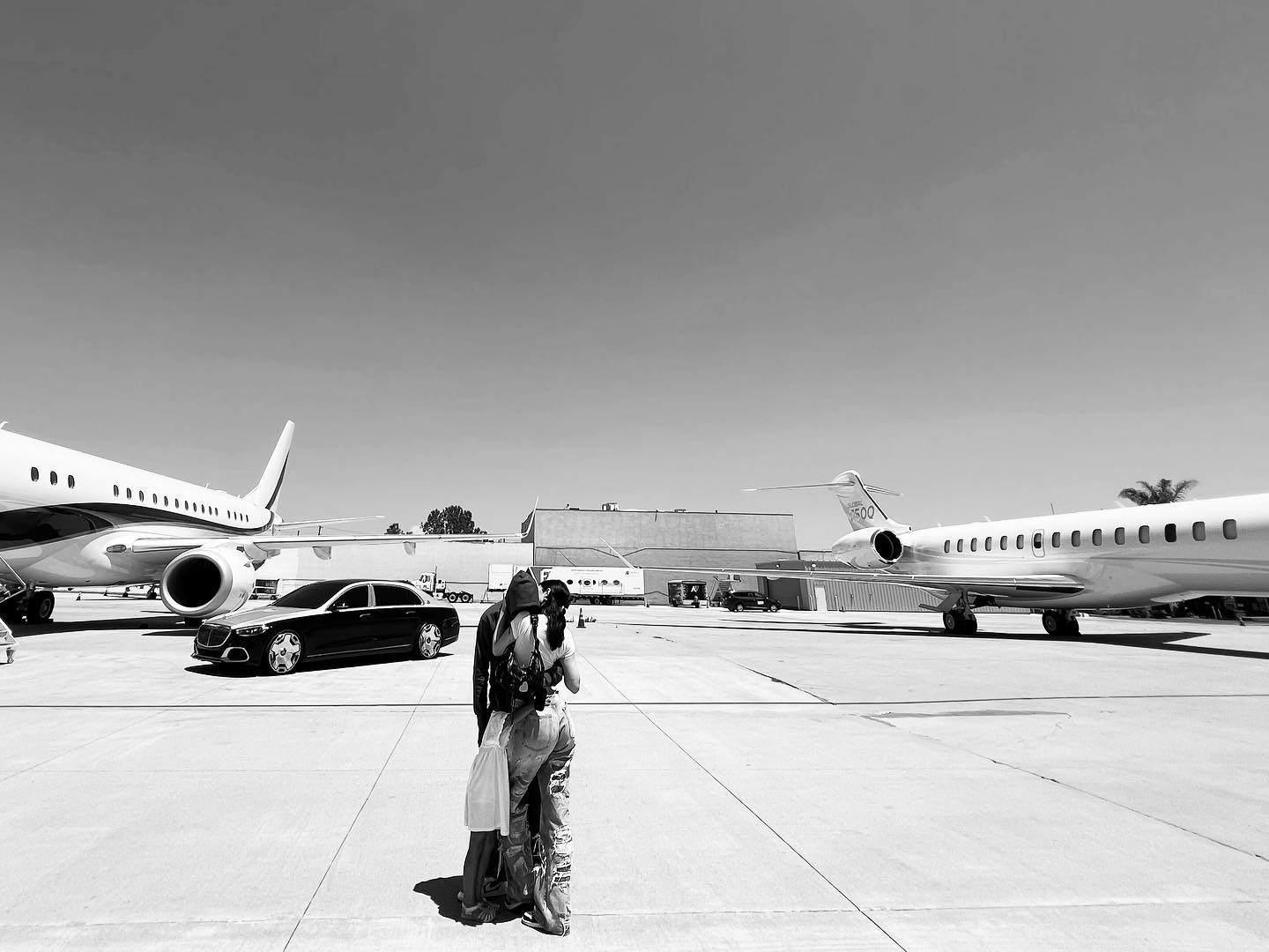 A couple embraces on an airport tarmac between luxury private jets and a black sedan, illustrating lavish travel lifestyle.