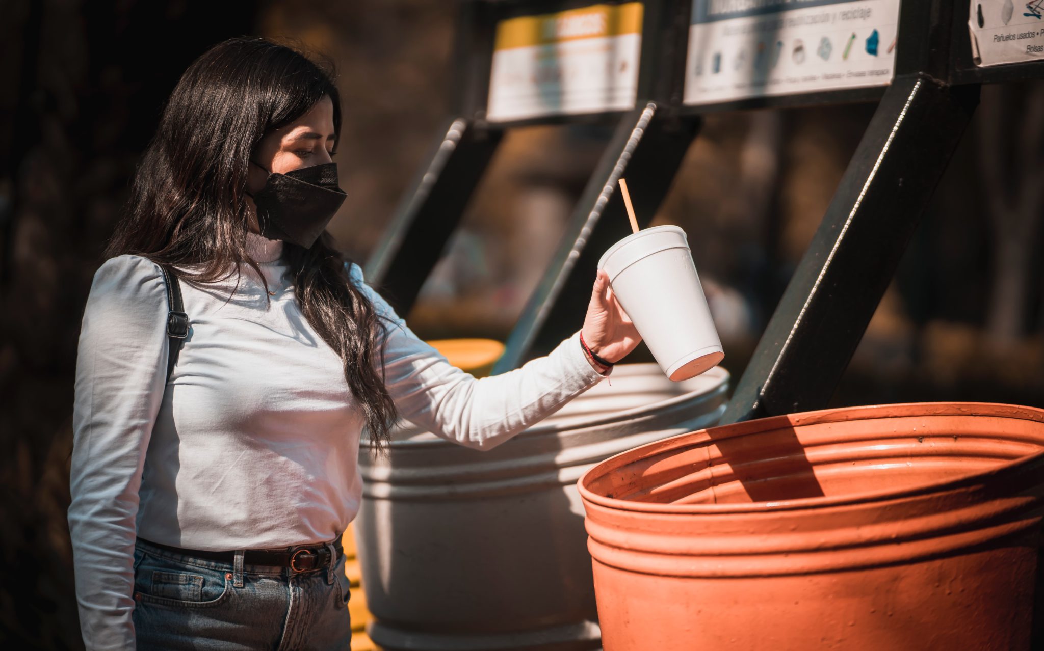 Woman wearing face mask disposes of white cup with paper straw into orange recycling bin at restaurant.