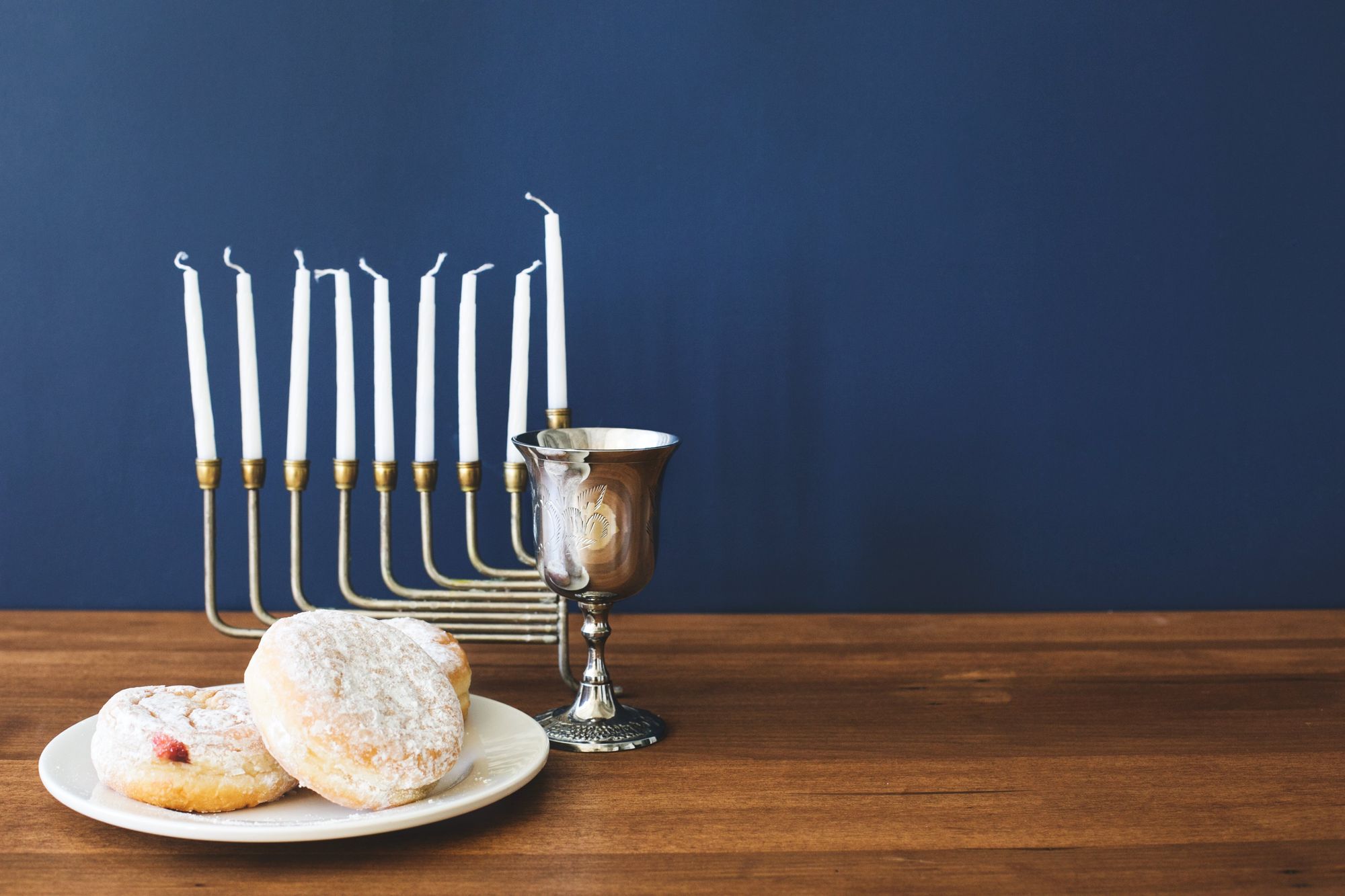 A menorah with white candles, silver kiddush cup, and plate of powdered sugar sufganiyot donuts on a wooden table.