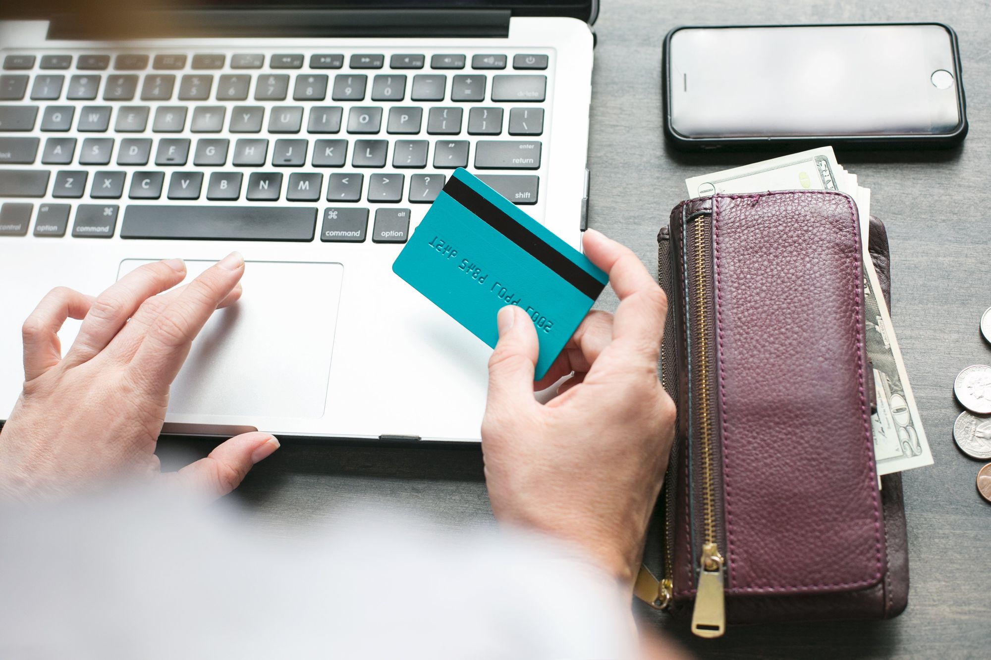 Person holding a teal credit card while using laptop, with wallet containing cash and smartphone nearby on desk.