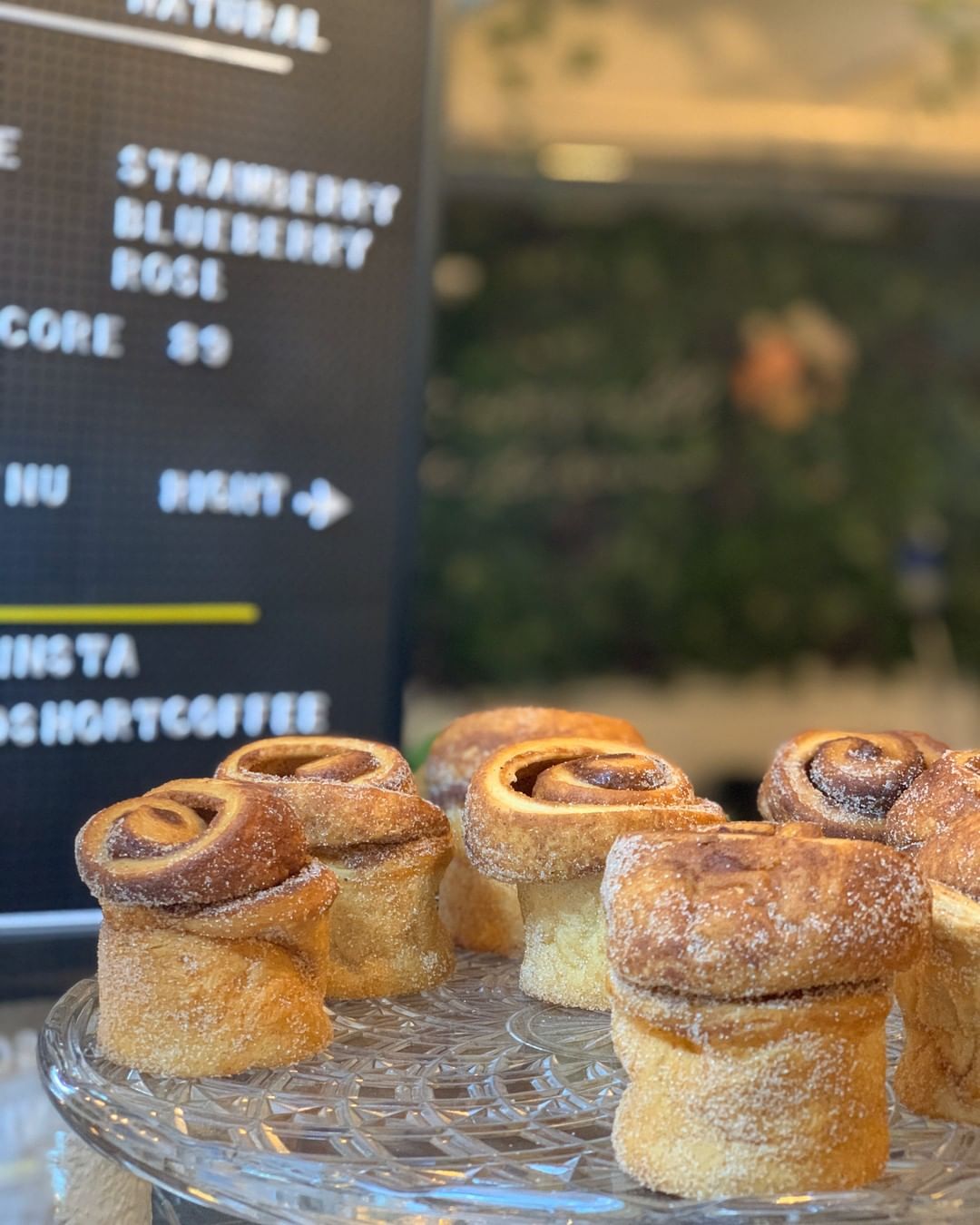 Golden cinnamon rolls with swirled tops displayed on a glass cake stand in a café, with a blurred menu board visible in the background.