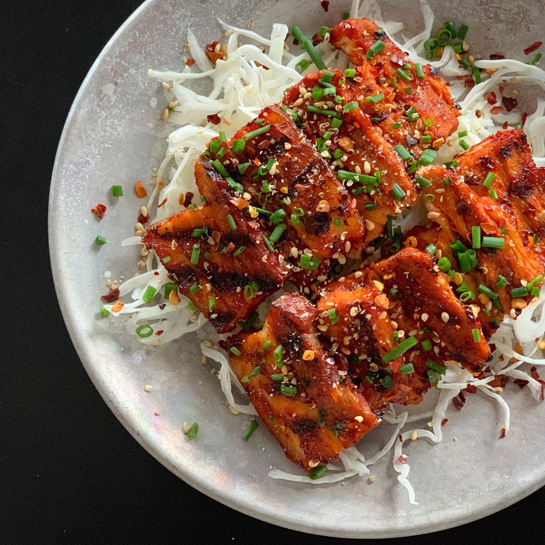 Grilled tofu glazed with spicy gochujang sauce, garnished with scallions and sesame seeds, served over rice noodles on a ceramic plate.