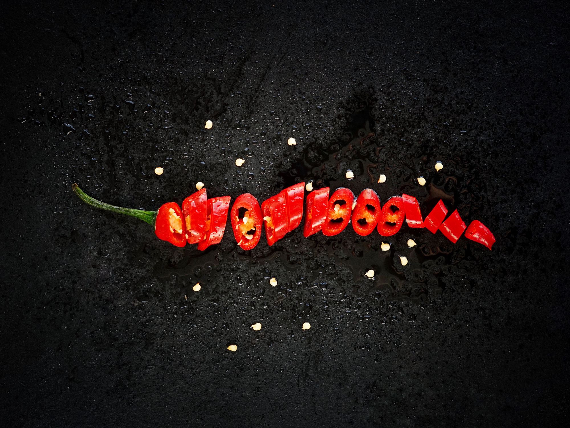 A sliced red chili pepper arranged on a dark surface with scattered garlic cloves and spices.