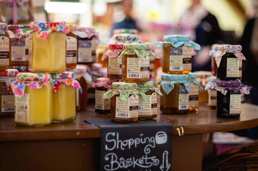 Artisanal jams and preserves with colorful fabric tops displayed on wooden shelves at a local farmers market.